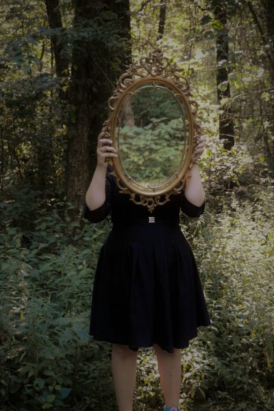 Femme dans un bois, un miroir cachant son visage et reflétant son image, symbolisant la réflexion personnelle et l’écriture de soi.
